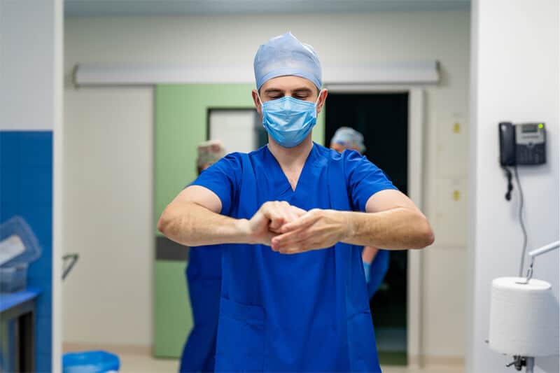 A surgeon in full blue scrubs, bouffant and face mask cleaning hands